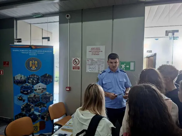 Un bărbat în uniformă albastră stă în picioare în fața unui grup de persoane care stau la mese într-o încăpere cu un panou informativ lateral.