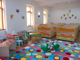 O cameră de joacă cu mese mici, scaune colorate, un covor cu modele circulare și paturi de copii aliniate pe partea dreaptă.
