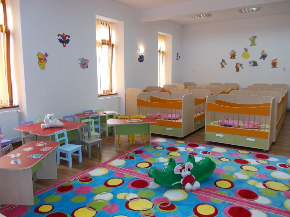 O cameră de joacă cu mese mici, scaune colorate, un covor cu modele circulare și paturi de copii aliniate pe partea dreaptă.