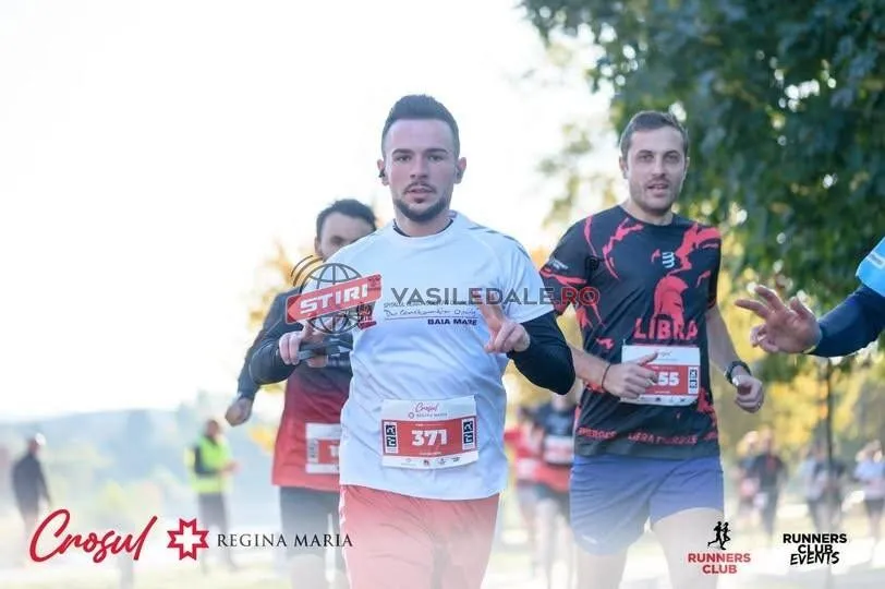 Un bărbat cu tricou alb și număr de concurs 371 aleargă pe o stradă alături de alți participanți la o cursă.