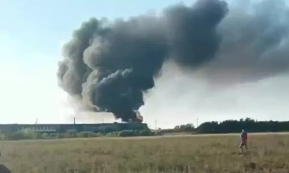 O coloană densă de fum negru se ridică deasupra unei clădiri joase situată în spatele unui câmp de iarbă uscată sub un cer deschis.