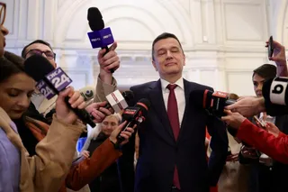 Un bărbat într-un costum închis și cravată roșie stă înconjurat de microfoane întinse de către mai mulți oameni.