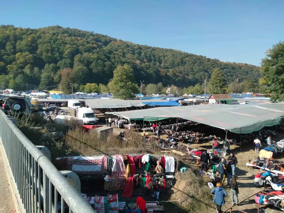 O vedere de sus asupra unui târg în aer liber cu tarabe acoperite, produse expuse pe pământ și oameni printre standuri, având un deal împădurit în fundal.