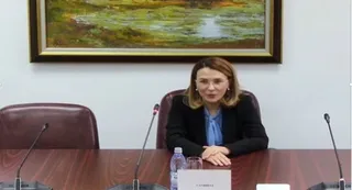 O femeie cu părul blond, purtând o jachetă neagră și o cămașă albastră, stă la o masă de conferință cu microfoane și o sticlă de apă.