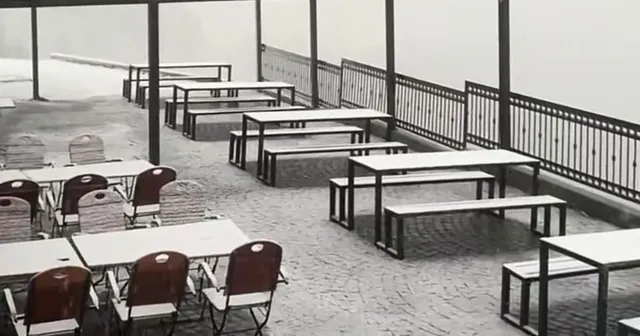 O terasă exterioară cu mese și bănci albe dispuse pe un pavaj, sub un cer gri și brumă.