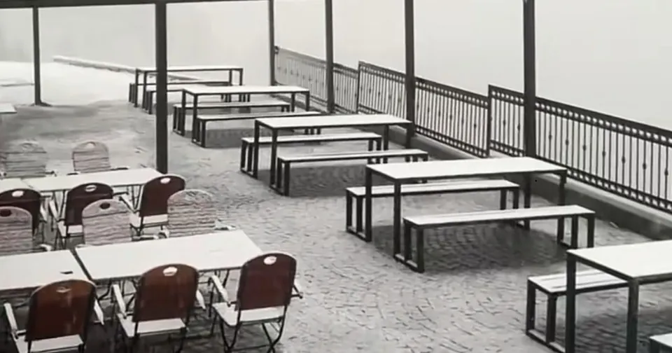 O terasă exterioară cu mese și bănci albe dispuse pe un pavaj, sub un cer gri și brumă.