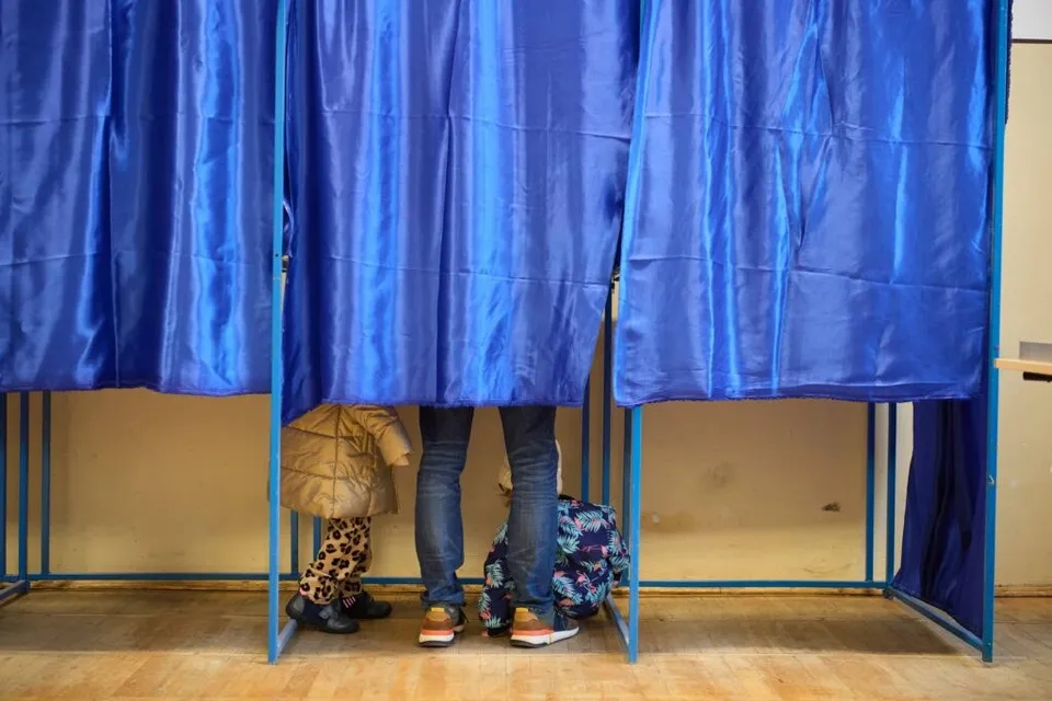 Două persoane stau în spatele unor cortine albastre de vot, fiind vizibile doar picioarele lor și partea inferioară a hainelor.