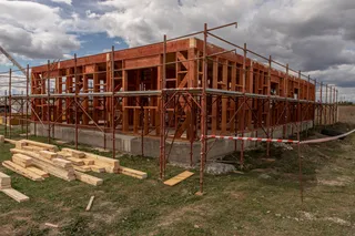 O structură de lemn pentru o clădire este înconjurată de niște schele metalice pe un teren cu iarbă și scânduri împrăștiate pe sol.