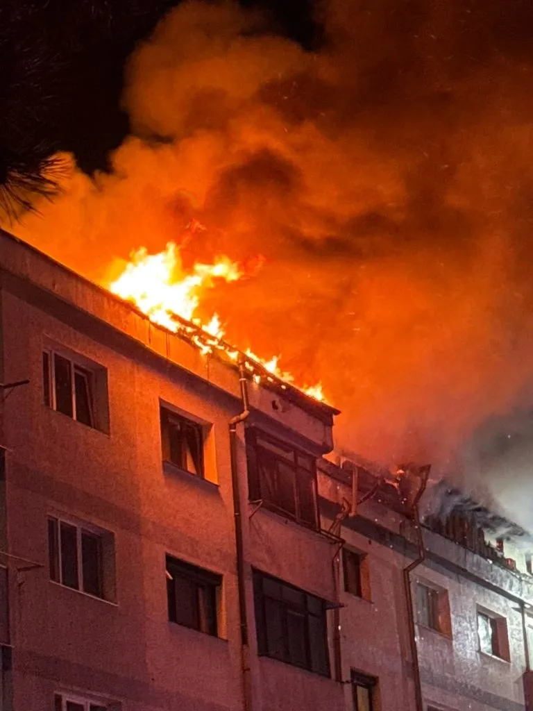 Flăcări mari și fum dens ies de pe acoperișul unei clădiri cu ferestre multiple pe timp de noapte.