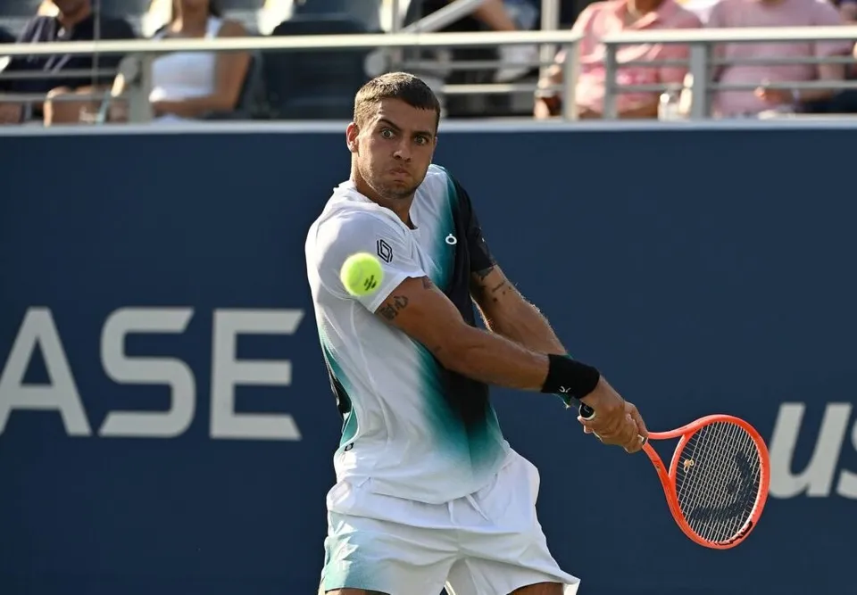 Un jucător de tenis în echipament alb și negru lovește o minge galbenă cu o rachetă portocalie pe un teren de sport.