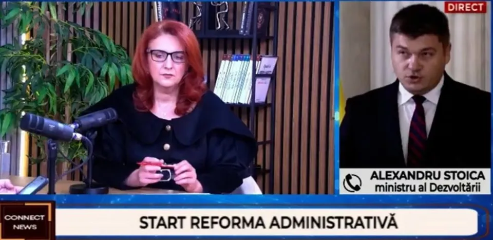 O femeie cu părul roșu și un bărbat în costum purtând cravată sunt prezentați într-un cadru de emisiune televizată.
