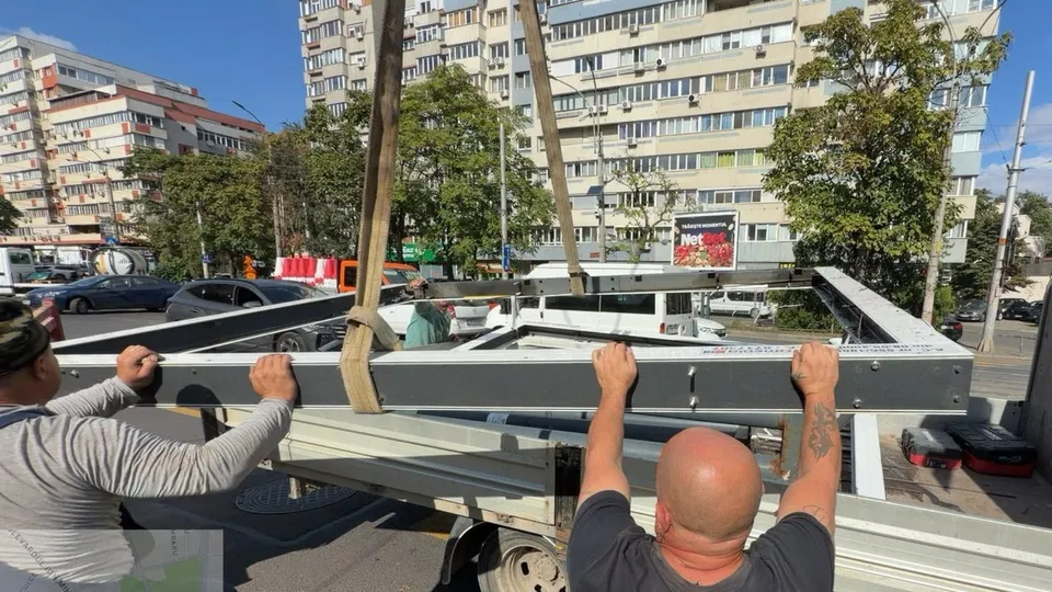 Două persoane ridică o structură metalică gri de pe un vehicul de transport aflat pe un drum urban lângă clădiri înalte.