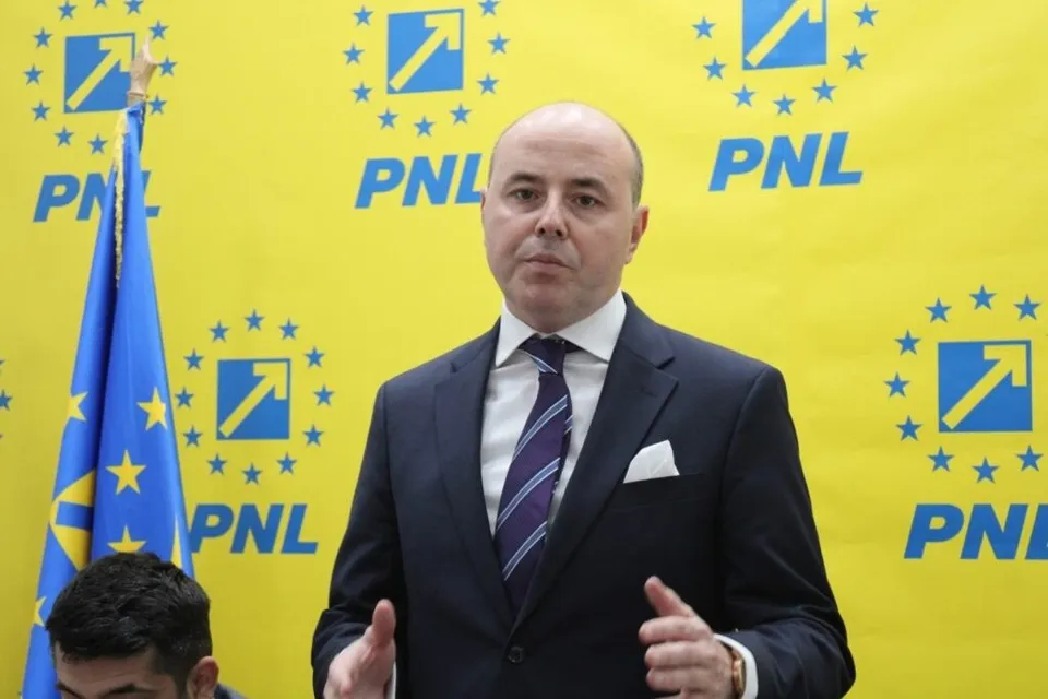 Un bărbat cu pielea deschisă, purtând sacou închis la culoare, cămașă albă și cravată cu dungi, stă în fața unui fundal galben cu logouri albastre.