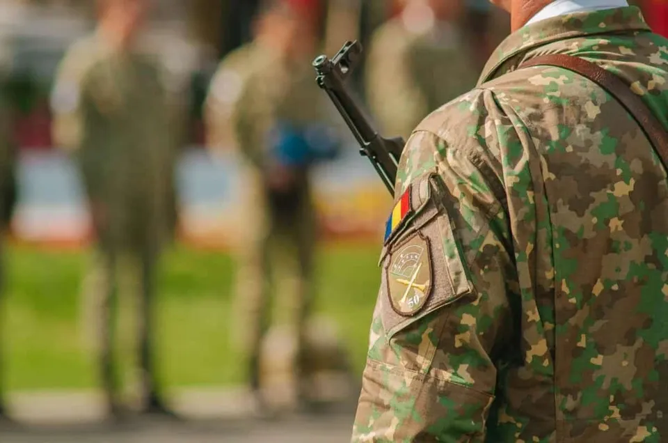 Un soldat în uniformă de camuflaj cu un emblema pe braț stă în picioare, având alți soldați desfocați în fundal.