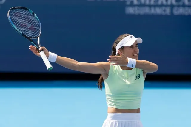 O jucătoare de tenis cu echipament alb și verde deschis, cu o vizoră albă, lovește mingea cu racheta într-o poziție extinsă pe un teren albastru.
