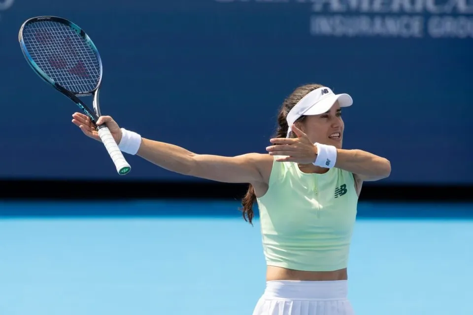 O jucătoare de tenis cu echipament alb și verde deschis, cu o vizoră albă, lovește mingea cu racheta într-o poziție extinsă pe un teren albastru.