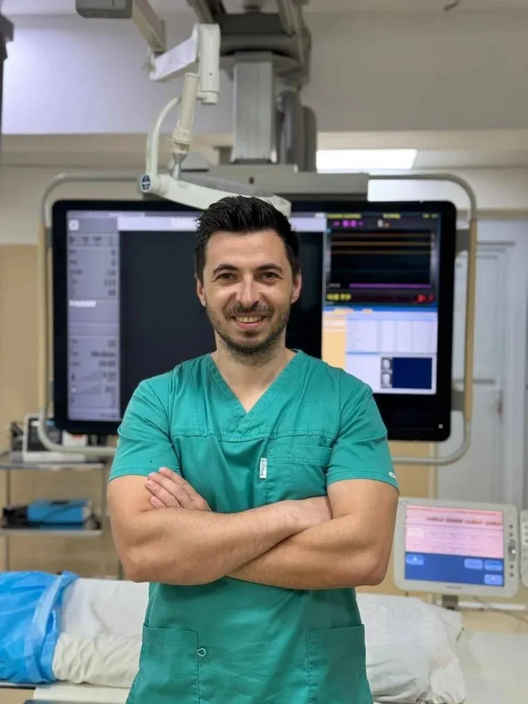 Un bărbat cu părul scurt și haine medicale verzi stă cu brațele încrucișate în fața unor monitoare medicale într-o încăpere iluminată.