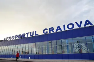 O persoană cu o vestă roșie merge pe trotuar în fața unei clădiri albastre cu ferestre mari și inscripția Aeroportul Craiova.