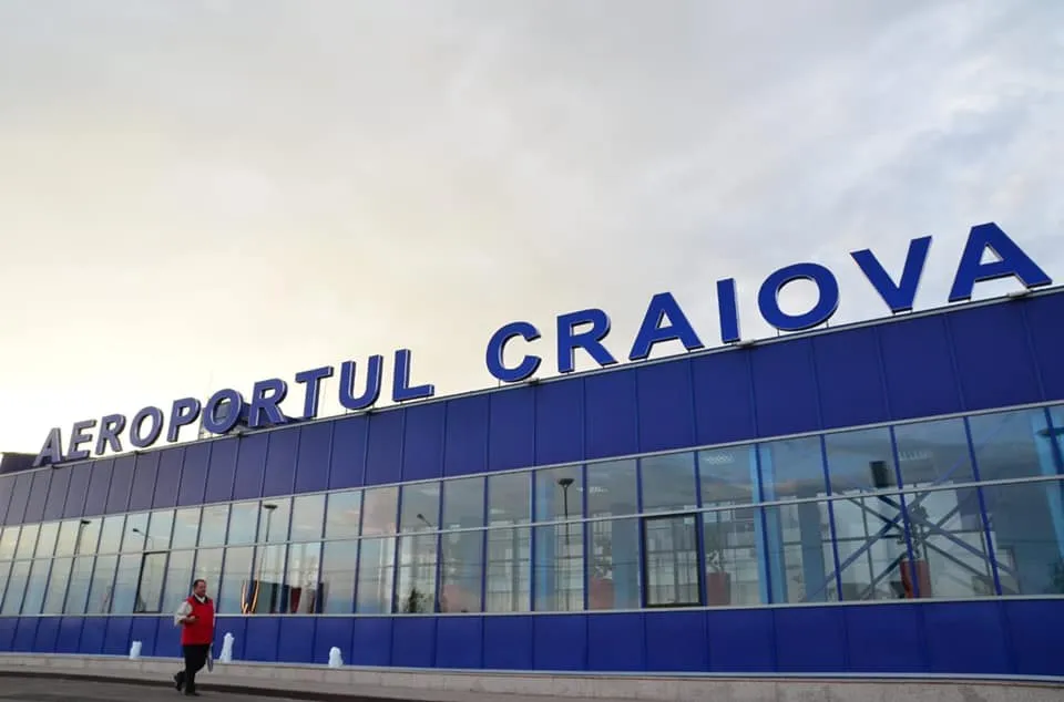 O persoană cu o vestă roșie merge pe trotuar în fața unei clădiri albastre cu ferestre mari și inscripția Aeroportul Craiova.