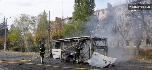 Două persoane cu echipament de protecție și cască stau lângă un autobuz distrus care scoate fum pe un drum asfaltat.