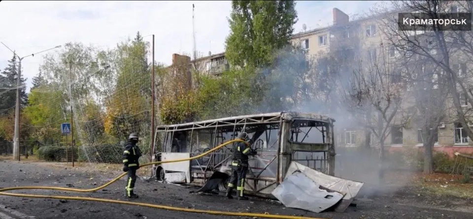 Două persoane cu echipament de protecție și cască stau lângă un autobuz distrus care scoate fum pe un drum asfaltat.