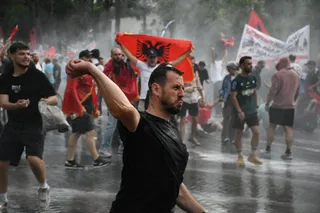 Un bărbat cu tricou negru stă în prim-plan, în timp ce mulțimea de oameni din spate poartă un steag roșu cu un vultur negru.