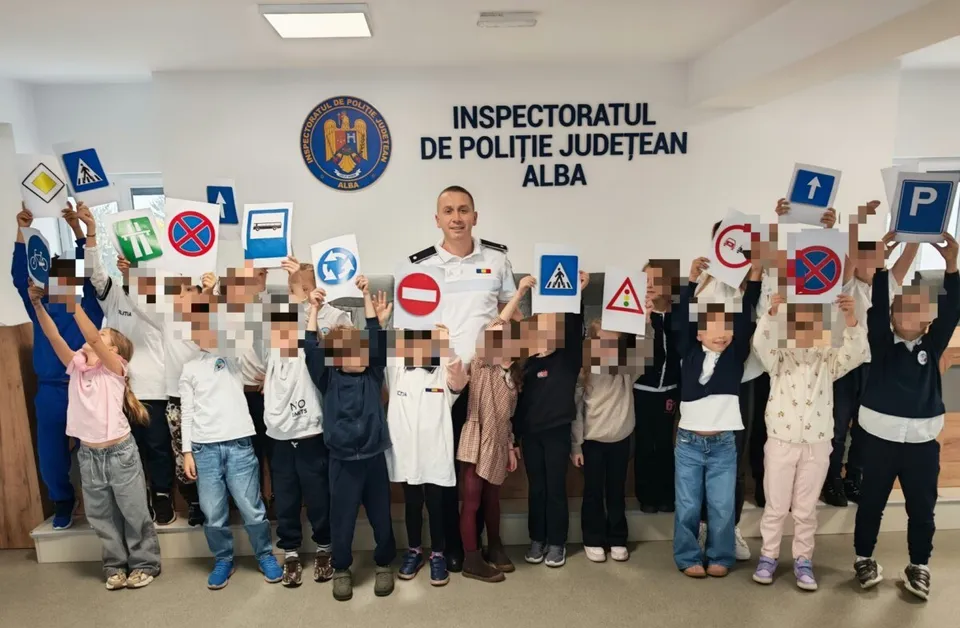 Un grup de copii și un polițist în uniformă albă stau în fața unui perete cu un emblema și un text, ținând diverse semne rutiere colorate.