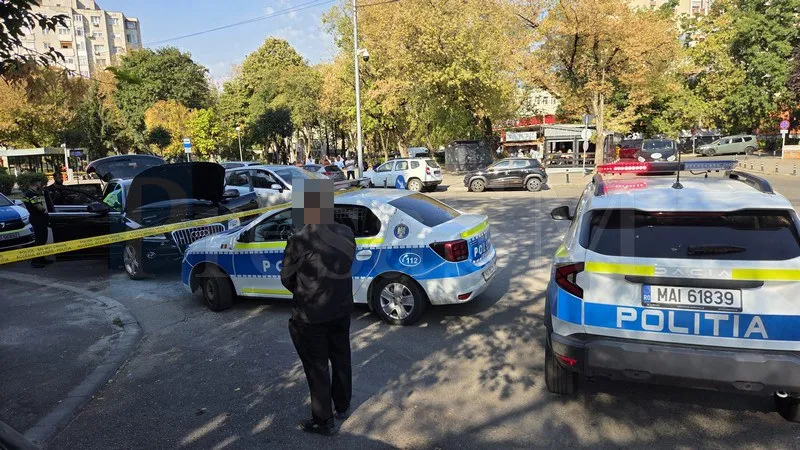 Două mașini de poliție și un vehicul alb sunt parcate pe un drum, lângă o bandă galbenă de delimitare și un bărbat cu spatele.
