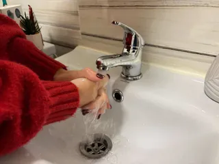 O persoană cu mâinile sub un robinet de la care curge apă într-o chiuvetă albă.