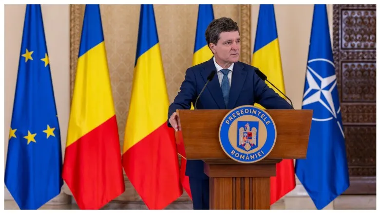 Un bărbat într-un costum închis la culoare și cravată stă în spatele unui podium de lemn în fața mai multor steaguri.