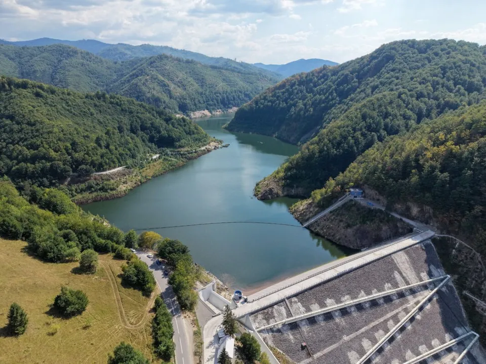 O vedere aeriană prezintă un lac înconjurat de dealuri împădurite și o parte dintr-un baraj de beton în prim-plan.