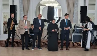 Șase persoane stau în picioare pe o scenă, un bărbat cu saxofon, un preot în haine negre și alți patru bărbați în costume.
