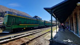 Un locomotor verde și locomotiva cu vagoane alăturate pe o cale ferată lângă o peron acoperit cu o structură de metal și o clădire de zidărie.