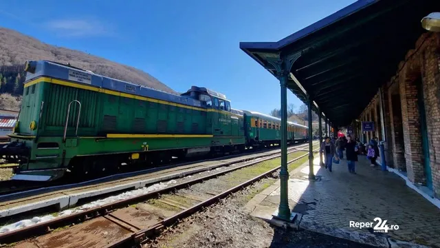 Un locomotor verde și locomotiva cu vagoane alăturate pe o cale ferată lângă o peron acoperit cu o structură de metal și o clădire de zidărie.