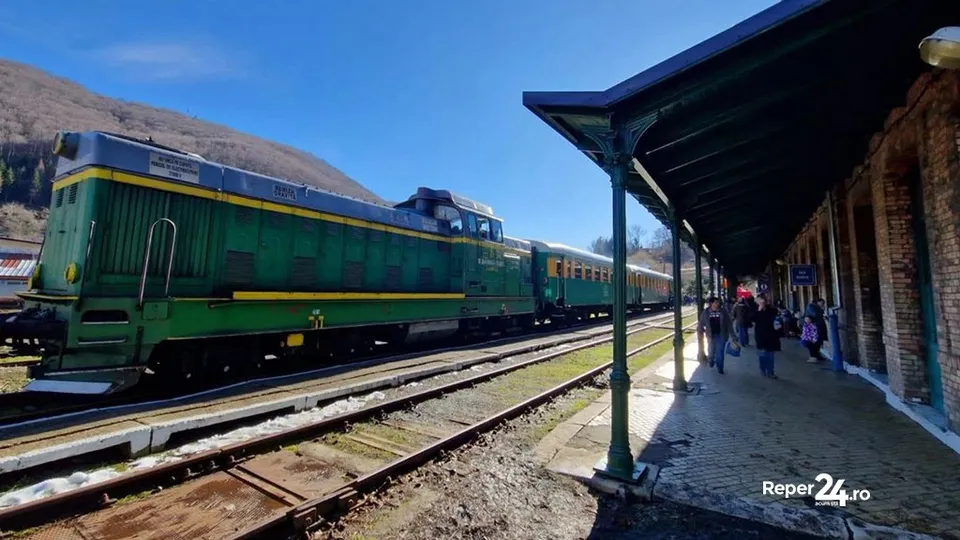 Un locomotor verde și locomotiva cu vagoane alăturate pe o cale ferată lângă o peron acoperit cu o structură de metal și o clădire de zidărie.