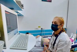 O femeie cu halat alb și mască medicală folosește un aparat medical lângă un pacient întins pe o masă de examinare.