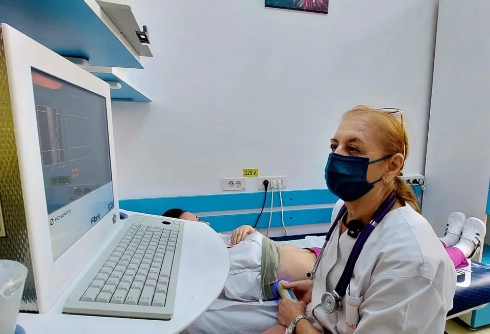 O femeie cu halat alb și mască medicală folosește un aparat medical lângă un pacient întins pe o masă de examinare.