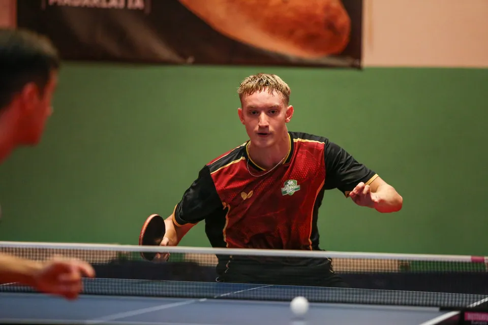 Un bărbat cu tricou negru și roșu ține o paletă de tenis de masă lângă o masă de joc cu o minge albă pe suprafață.