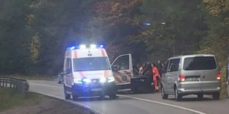 O ambulanță cu girofaruri aprinse stă pe marginea unui drum asfaltat lângă un autoturism și o mașină de serviciu cu portierele deschise.