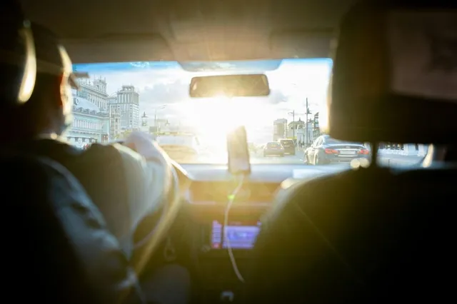 Persoana conduce un autovehicul pe un drum urban, fiind vizibilă perspectiva din interiorul mașinii spre parbriz și traficul exterior sub lumina soarelui.