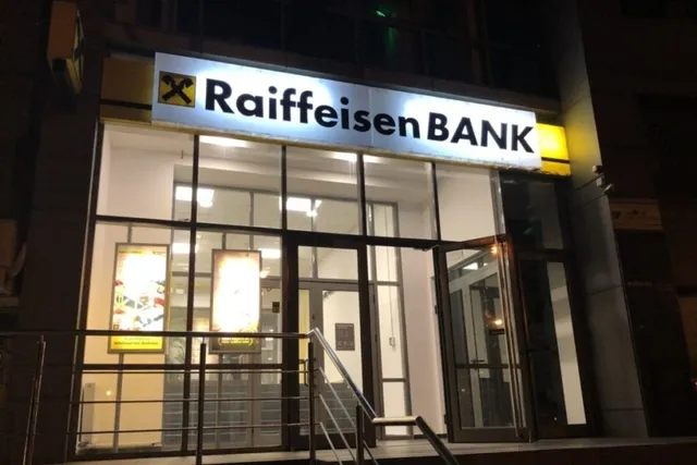 O fațadă de clădire iluminată noaptea prezintă un panou cu inscripția Raiffeisen BANK deasupra unor uși și ferestre de sticlă.