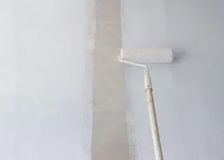 Un rulou de vopsit cu mâner lung aplică o substanță de culoare deschisă pe o suprafață verticală gri pală.