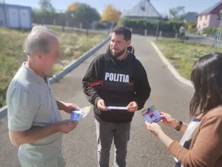 Un bărbat cu hanorac negru oferă pliante unor două persoane aflate pe un drum asfaltat.