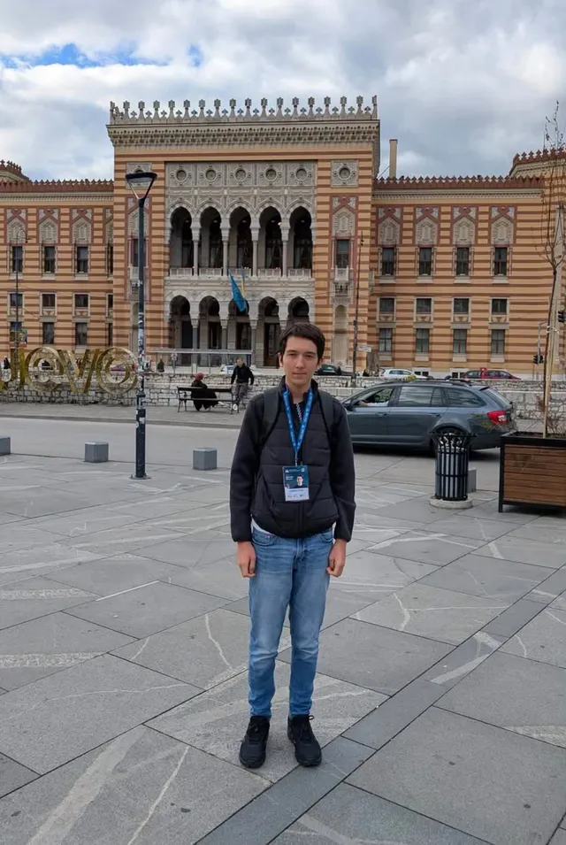 Un tânăr cu sacou negru și blugi deschizi stă în fața unei clădiri mari cu ferestre arcuite și un pavaj de piatră.