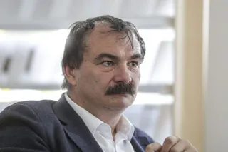Bustul unui bărbat cu mustață, în sacou și cămașă, privind lateral, cu o mână parțial vizibilă.