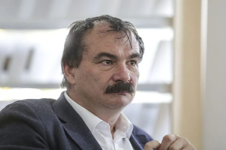 Bustul unui bărbat cu mustață, în sacou și cămașă, privind lateral, cu o mână parțial vizibilă.