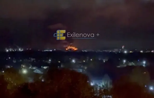 O lumină portocalie strălucitoare este vizibilă pe orizontul unei zone urbane întunecate sub un cer nocturn.