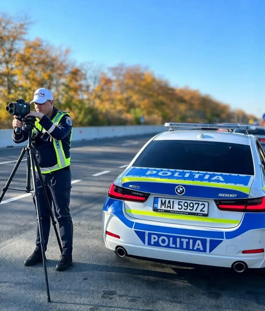 Un polițist cu vestă reflectorizantă folosește o cameră video montată pe un trepied lângă o mașină de poliție albă, albastră și galbenă.