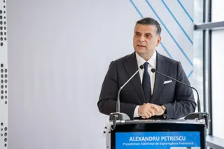 Un bărbat într-un costum închis la culoare și cravată stă în spatele unui podium cu microfoane.
