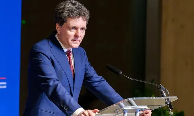 Un bărbat cu părul închis la culoare, îmbrăcat într-un costum albastru și cravată roșie, stă în spatele unui podium transparent cu un microfon.
